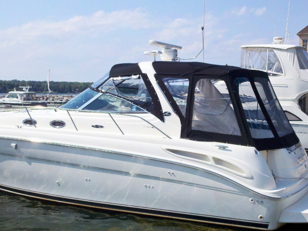 The side of a large white boat with a black canvas enclosure that has clear vinyl windows. Another boat with a similar clear enclosure is visible in the background.