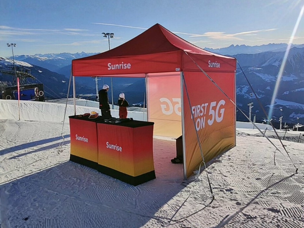 A red and orange branded promotional booth or canopy tent, labeled "Sunrise" and "FIRST ON 5G," set up on a snowy mountain slope. Two people are standing near the counter, with ski lift infrastructure and distant snow-capped mountains in the background.