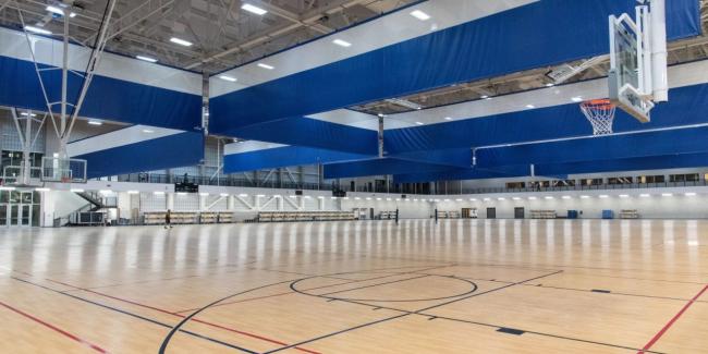 A spacious indoor gymnasium with a polished wooden floor, multiple basketball hoops, and large blue and white retractable divider curtains hanging from the ceiling